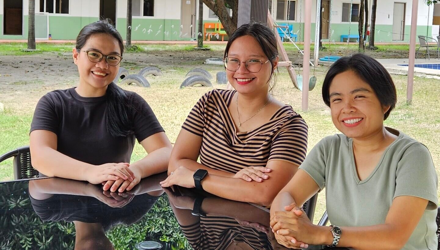 three staff of BELA E4U Academy are smiling and seated. They are surrounded by dormitories of BELA Academy.