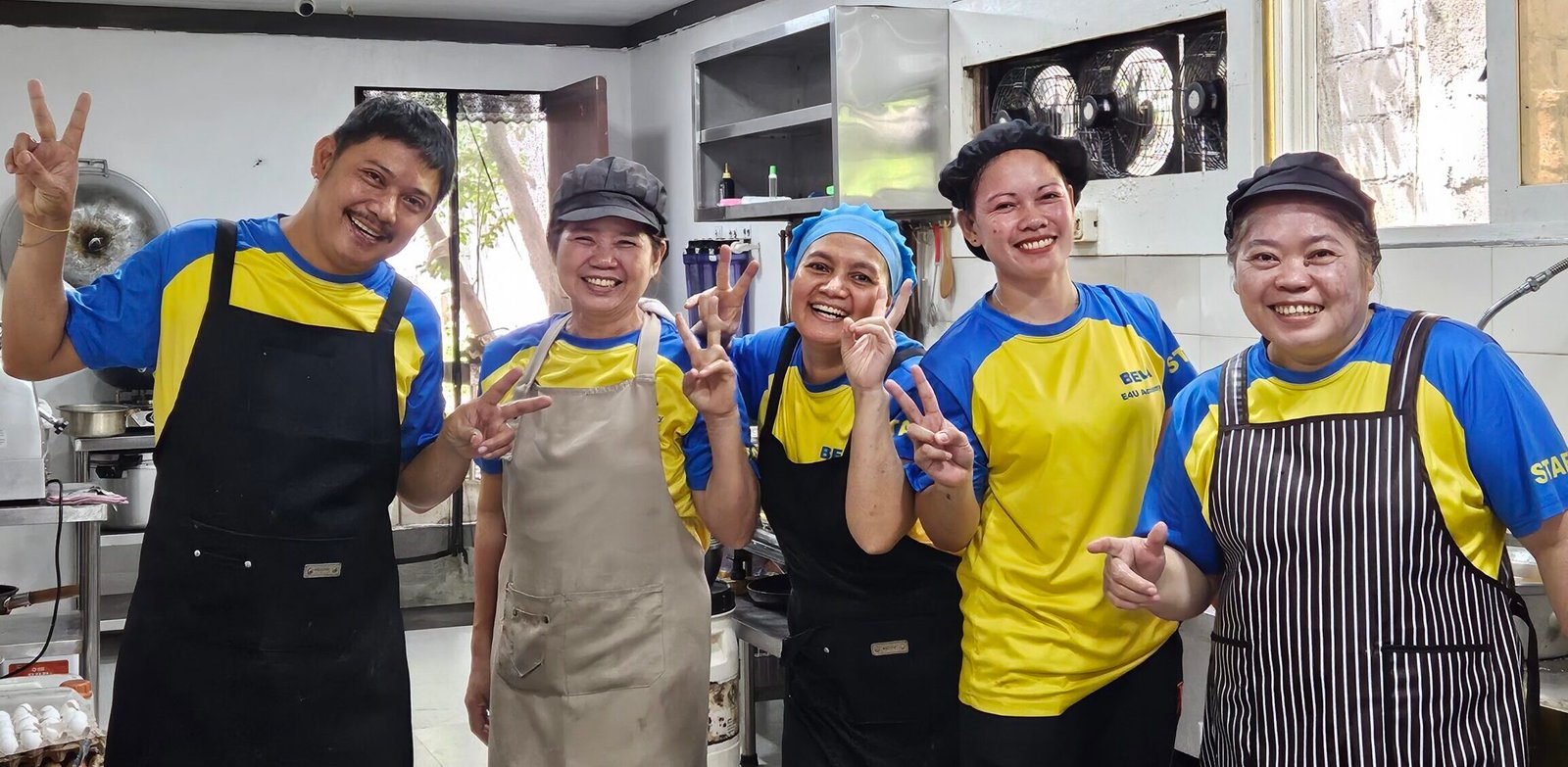 Five kitchen staff of BELA E4U Academy in the kitchen wearing aprons and headdress are waving and smiling.